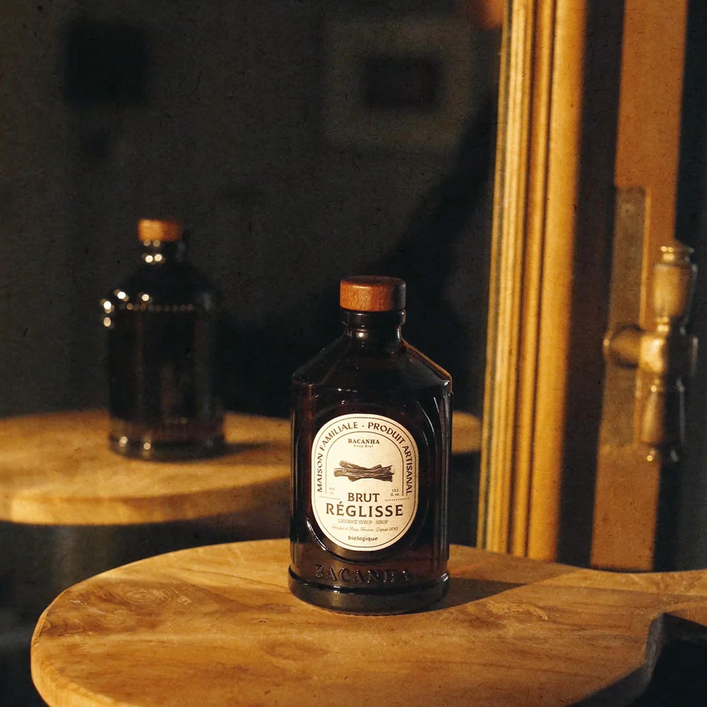 Bacanha Bio-Lakritz-Sirup (Sirop Brut de Réglisse) in der 400ml Design-Glasflasche neben einem Glas Eistee mit Strohhalm auf einem Holztisch.