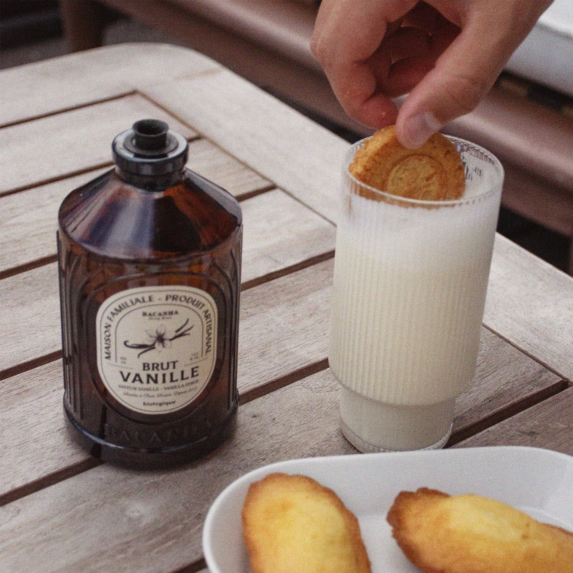 Bacanha Bio-Vanille-Sirup (Sirop Brut de Vanille) in der 400ml Glasflasche präsentiert auf einem Holztisch neben einer Tasse Cappuccino – ideal zum Verfeinern von Kaffee und Desserts.