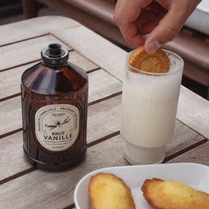 Bacanha Bio-Vanille-Sirup (Sirop Brut de Vanille) in der 400ml Glasflasche präsentiert auf einem Holztisch neben einer Tasse Cappuccino – ideal zum Verfeinern von Kaffee und Desserts.