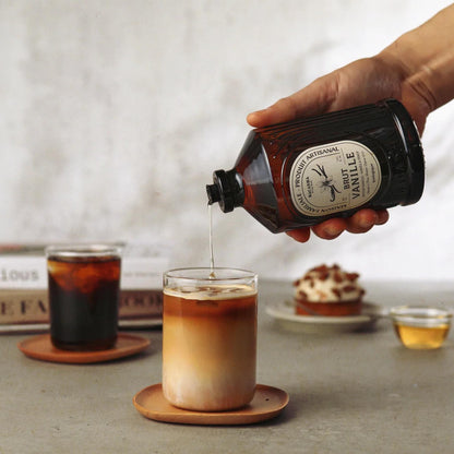Bacanha Bio-Vanille-Sirup (Sirop Brut de Vanille) wird aus der 400ml Glasflasche in eine Tasse Kaffee mit Milchschaum gegossen – hochwertiger Bio-Sirup aus Frankreich zur Verfeinerung von Kaffeespezialitäten.
