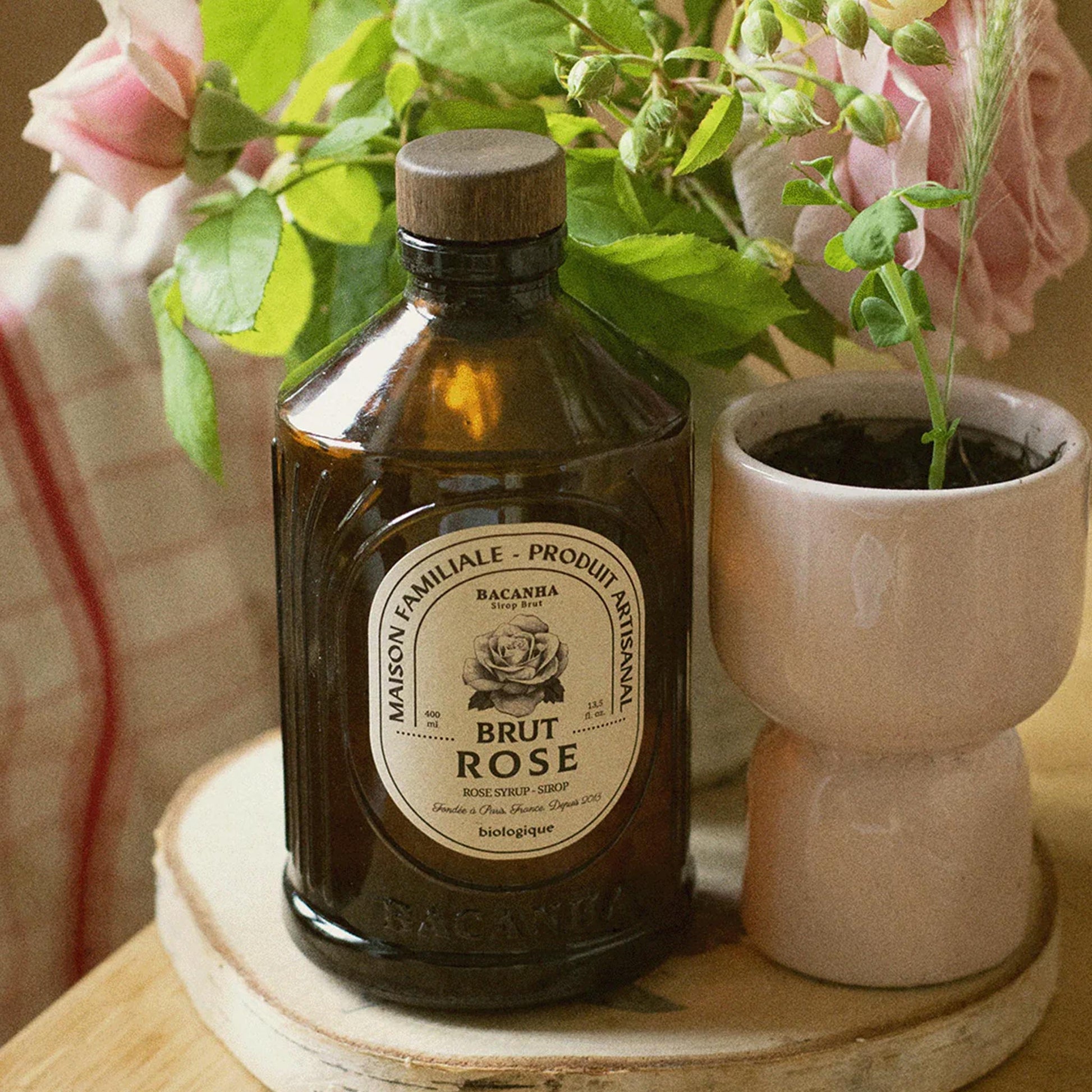 Bacanha Bio-Rosensirup (Sirop Brut de Rose) in der 400ml Glasflasche neben einem Glas mit Eiswürfeln, einer Rosenblüte und einem Strohhalm auf einem rustikalen Holztisch.