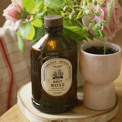 Bacanha Bio-Rosensirup (Sirop Brut de Rose) in der 400ml Glasflasche neben einem Glas mit Eiswürfeln, einer Rosenblüte und einem Strohhalm auf einem rustikalen Holztisch.