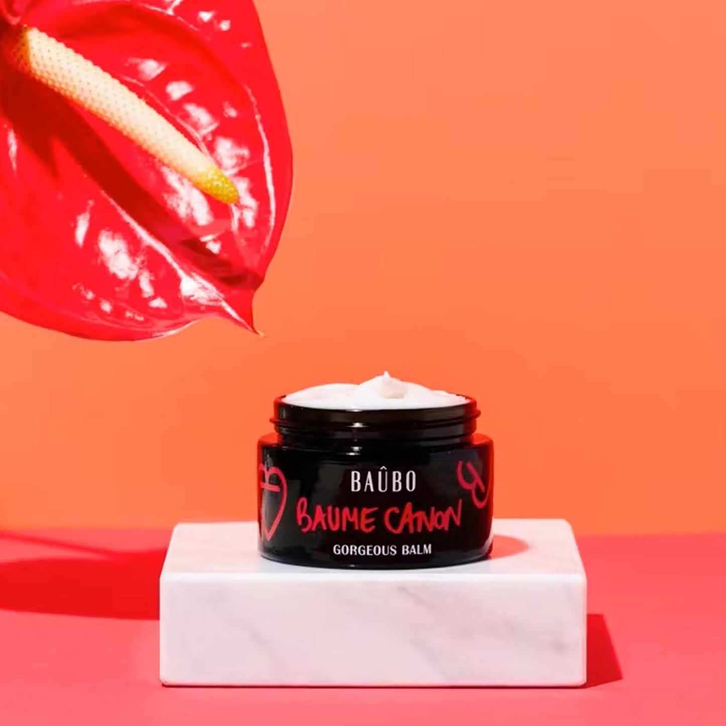 Geöffneter Tiegel Baûbo Baume Canon Gorgeous Balm (50 ml) mit cremiger Bio-Textur, präsentiert auf einem Marmorpodest vor korallenfarbenem Hintergrund.