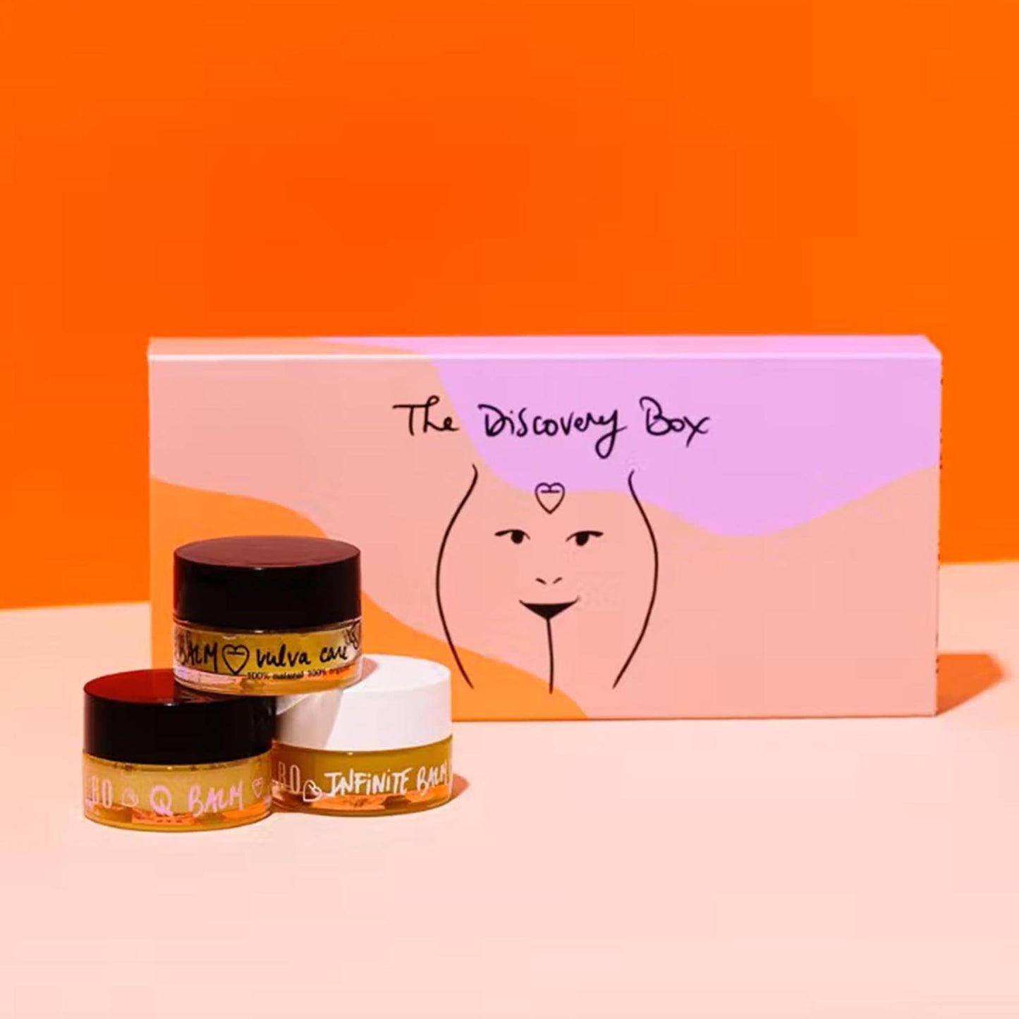Baûbo „The Discovery Box“ Geschenkset (3 x 15 ml) – Drei Bio-Pflegebalsame (Vulva Care, Q Balm, Infinite Balm) vor einer illustrierten Verpackung auf orangefarbenem Hintergrund.