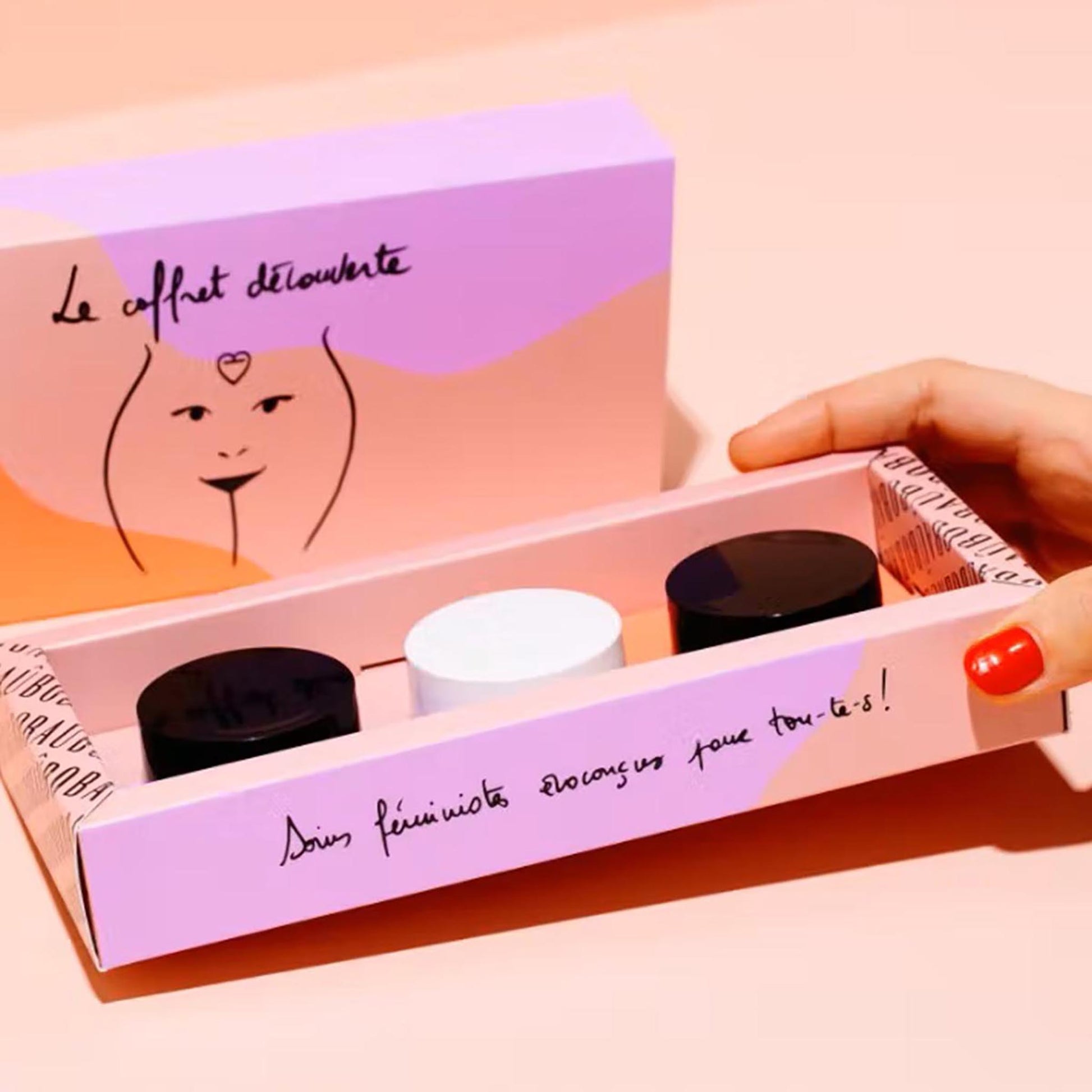 Geöffnete Baûbo „The Discovery Box“ (3 x 15 ml) mit drei Bio-Pflegebalsamen in einer rosa Geschenkverpackung, gehalten von einer Hand mit rot lackierten Fingernägeln.