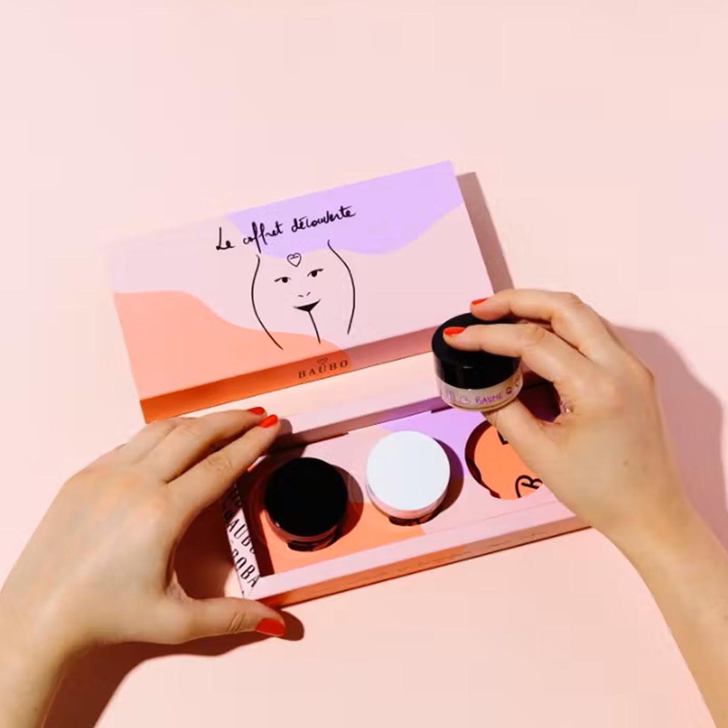 Draufsicht auf die geöffnete Baûbo „The Discovery Box“ (3 x 15 ml) – Hände mit rot lackierten Fingernägeln entnehmen einen der drei Bio-Pflegebalsame aus dem farbenfrohen Geschenkset.