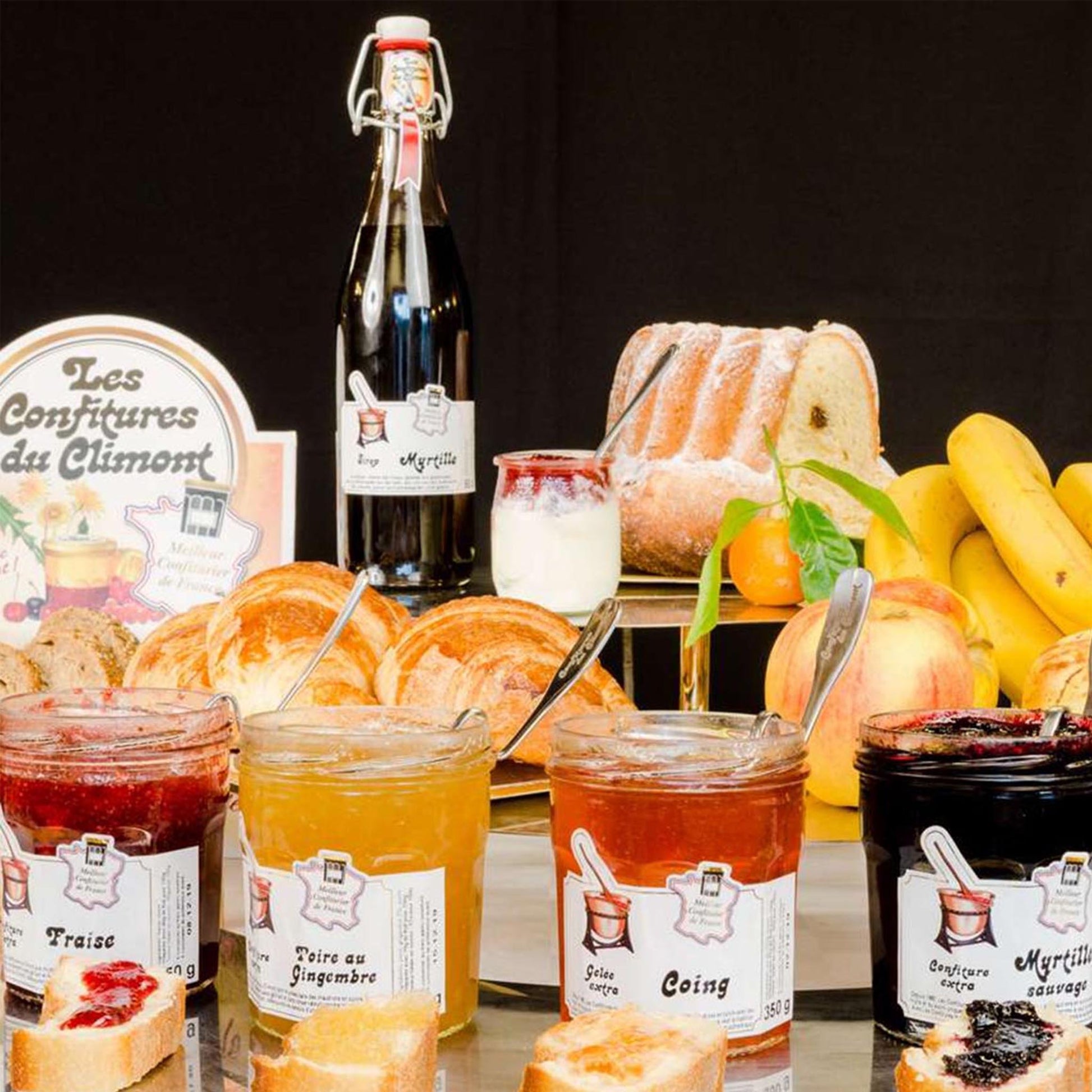 Auswahl handwerklich hergestellter Konfitüren von Les Confitures du Climont mit Erdbeere, Birne mit Ingwer, Quitte und wilder Heidelbeere, arrangiert mit Brot, Croissants, Gugelhupf, Früchten und Flasche auf gedecktem Tisch.