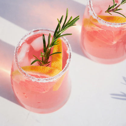 Erfrischender Cocktail mit Bacanha Sirop Brut de Romarin Bio – rosafarbener Drink mit Rosmarinzweig, Zitrusschale und Zuckerrand, serviert auf Eis