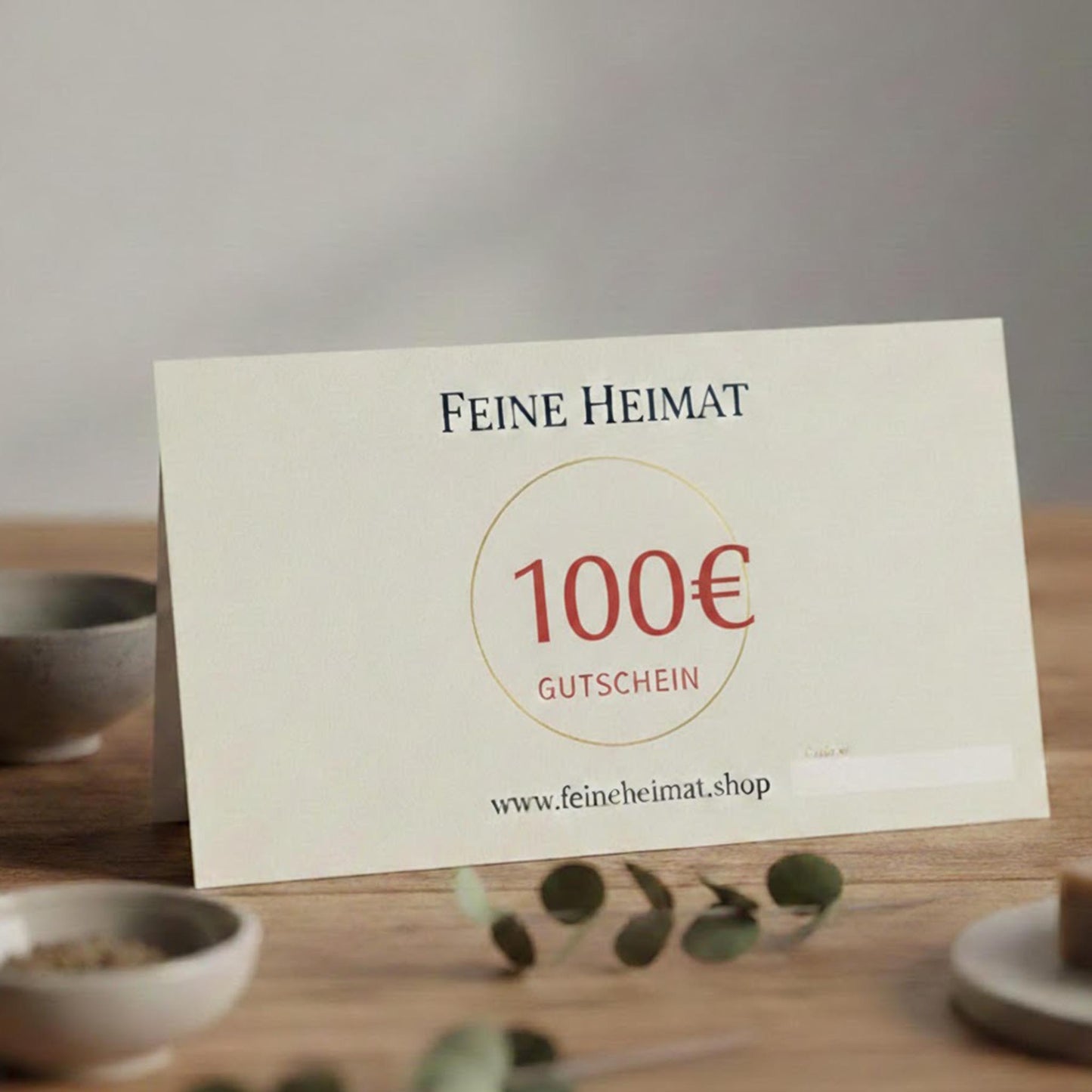 100 Euro Geschenkgutschein von Feine Heimat – Hochwertige Gutscheinkarte auf einem Holztisch, ansprechend dekoriert mit Eukalyptus und kleinen Schalen.
