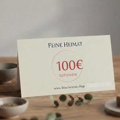 100 Euro Geschenkgutschein von Feine Heimat – Hochwertige Gutscheinkarte auf einem Holztisch, ansprechend dekoriert mit Eukalyptus und kleinen Schalen.