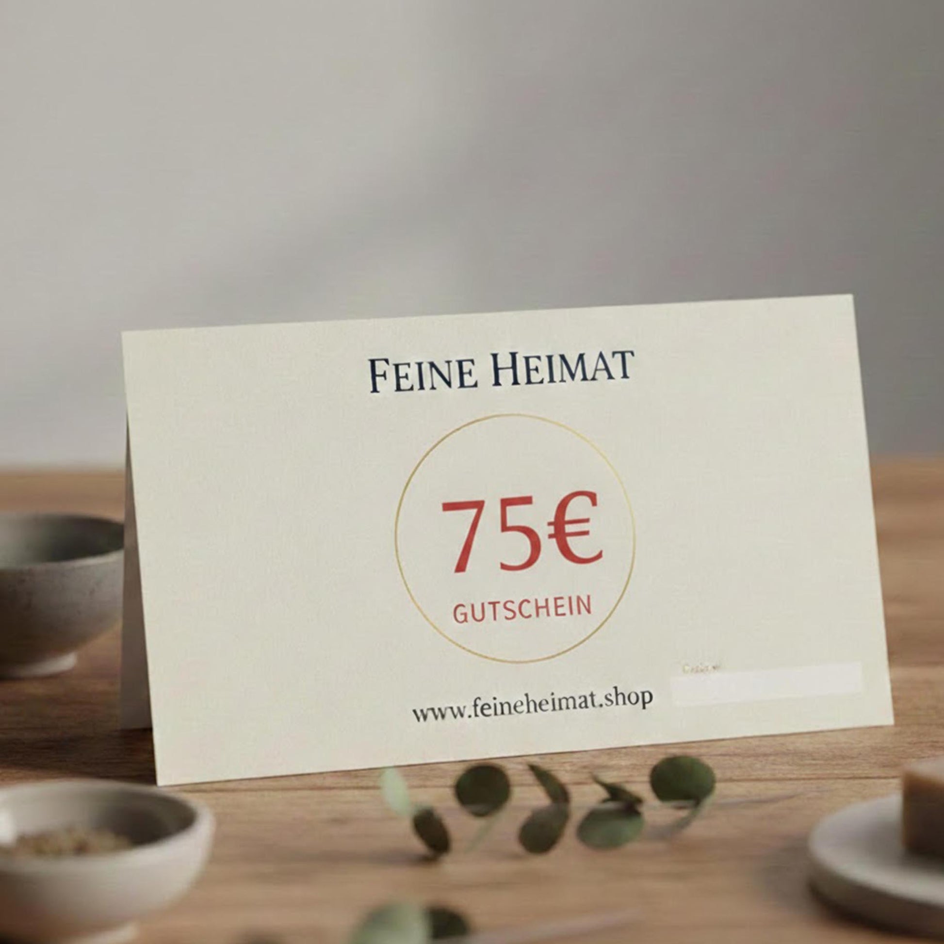 75 Euro Geschenkgutschein von Feine Heimat – Hochwertige Gutscheinkarte auf einem Holztisch, ansprechend dekoriert mit Eukalyptus und kleinen Schalen.
