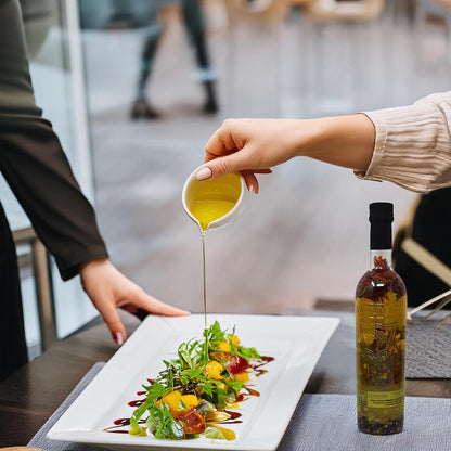 Lifestyle-Produktbild: Eine 500-ml-Glasflasche A L'Olivier Olivenöl mit Piment d’Espelette und Chili steht auf einem gedeckten Tisch, während im Hintergrund ein frischer Salat mit dem würzigen Öl verfeinert wird.